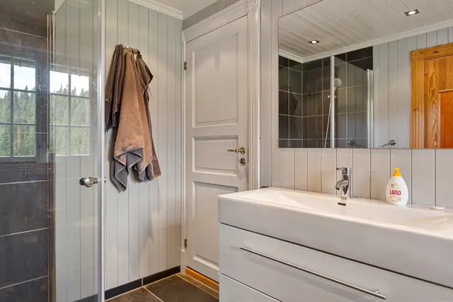Badet har mørke 30x60 cm fliser på gulv, med 30x30 cm fliser i det nedsenkede dusjområdet og veggene er kledd med mørke 30x30 cm fliser kombinert med malt panel.