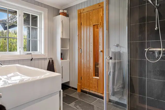 Badet er utstyrt med toalett og dusjhjørne med glassdører og ettgreps dusjarmatur. Baderomsinnredningen består av høyskap og 100 cm servantskap med glatte hvite fronter, servant og ettgreps krummet armatur.